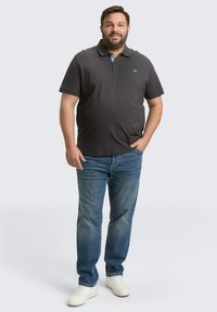 Mężczyzna z brodą ubrany w ciemnoszary polo, niebieskie dżinsy i białe trampki, stojący z jedną ręką w kieszeni na jednolitym tle.