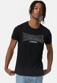 Melnas kokvilnas T-krekls ar baltu "LONSDALE" logotipu un "LONDON" tekstu. Īsas piedurknes un apaļš kakls, pieguļoša siluets.