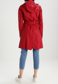 Wodoodporna czerwona peleryna przeciwdeszczowa z kapturem, paskiem w talii i lekko rozkloszowanym dołem. Połączona z przyciętymi niebieskimi dżinsami i białymi trampkami.