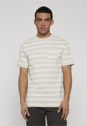 Młody mężczyzna o krótkich kręconych włosach, ubrany w beżowo-białą koszulę w paski z krótkim rękawem, z kieszenią na piersi, oraz w ciemne spodnie.