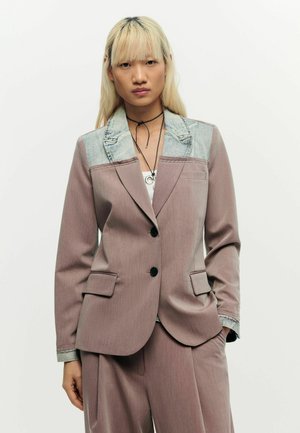 Ung person med langt blondt hår iført en mauve jakkesætjakke med denim-krave og matchende vide bukser, stående mod en hvid baggrund.