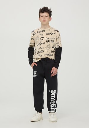 Felpa beige a maniche lunghe con accenti neri, caratterizzata da vari grafica stampate in nero. Pantaloni sportivi neri con testo e logo "Screamin" in bianco.