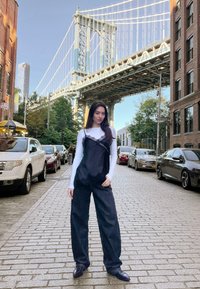 Giovane donna in tuta nera e camicia a maniche lunghe bianca che si trova su una strada di ciottoli, con un ponte e auto in sosta sullo sfondo.