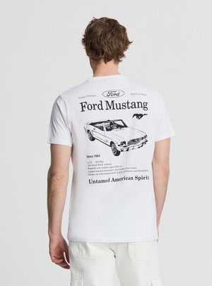 Mężczyzna w białej koszulce z czarną grafiką Ford Mustang oraz tekstem opisującym specyfikacje samochodu i hasłem „Nieokiełznany amerykański duch”.
