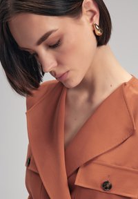 Burnt Orange Blazer mit strukturiertem Kragen und sichtbarem Knopfdetail. Der glatte Stoff und die scharfen Linien betonen das maßgeschneiderte Design. Goldene Tropfenohrringe.