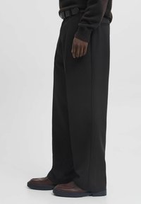 Czarne spodnie o szerokich nogawkach wykonane z gładkiego materiału, z przednią kieszenią i fakturowanym czarnym paskiem. Połączone z brązowym obuwiem.