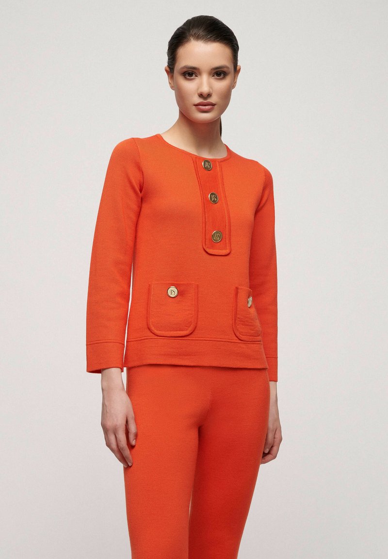 Oranges gestricktes Zweiteiler-Set mit einem langärmeligen Oberteil, das über einen runden Ausschnitt, vier goldene Knöpfe und zwei Fronttaschen verfügt. Die Hose ist eng anliegend.