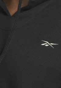 Detailní záběr na černou látku mikiny s kapucí, na které je bílý vyšívaný logotyp Reebok na hrudi a část šňůrky je viditelná.