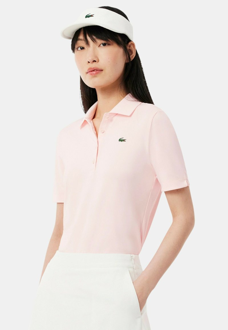 Polo shirt rosa chiaro con maniche corte, collo a polo e logo di coccodrillo ricamato verde. Abbinato a un visiera bianca.