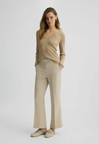 Maglione beige a costine con maniche lunghe e scollatura a V, abbinato a pantaloni beige chiaro a vita alta e mocassini color crema con un accento dorato.