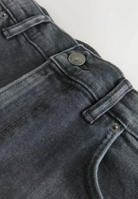 Primer plano de la cintura de unos vaqueros de mezclilla gris oscuro con pasadores para cinturón, botón de metal y costuras del bolsillo delantero sobre un fondo claro.