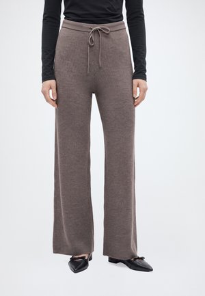 Un modèle porte un pantalon large côtelé couleur taupe à taille élastiquée avec un cordon de serrage, un haut noir et des chaussures plates noires, sur un fond blanc uni.