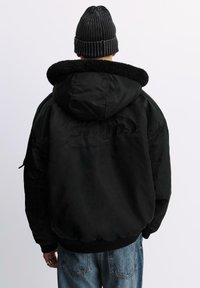 Schwarze Steppjacke mit einem fellgefütterten Kapuzen, mit einer glatten Außenseite und gerippten Bündchen. Auffälliges geprägtes Logo auf der Rückseite. Schwarze, gerippte Mütze.