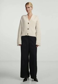 Ελεφαντίσια καγιούρκα με V-λαιμό και φαρδιές μανίκες, διακοσμημένη με τρία καφέ κουμπιά. Συνδυάζεται με μαύρα φαρδιά παντελόνια και μυτερά παπούτσια.