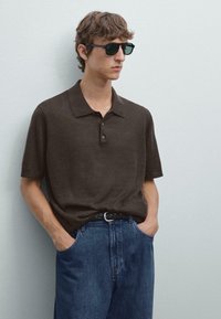 Hnědá polokošile s krátkým rukávem a límečkem, se třemi knoflíky, kombinovaná s modrými džíny s bočními kapsami. Na sobě má černé sluneční brýle.