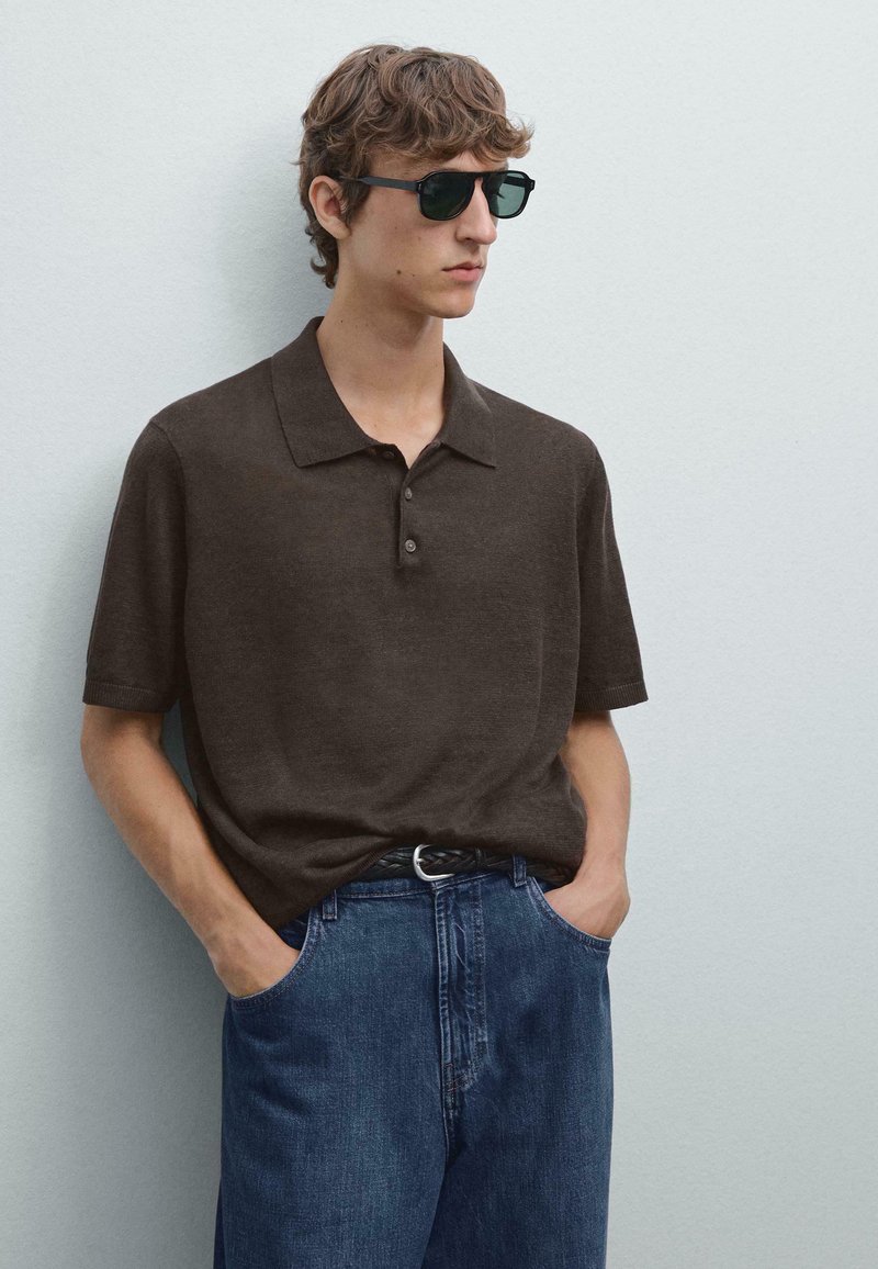 Hnědá polokošile s krátkým rukávem a límečkem, se třemi knoflíky, kombinovaná s modrými džíny s bočními kapsami. Na sobě má černé sluneční brýle.