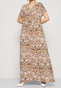 Robe maxi avec des manches courtes, décolleté en V et taille cintrée. Présente un motif géométrique multicolore sur un tissu léger. Talons blancs.