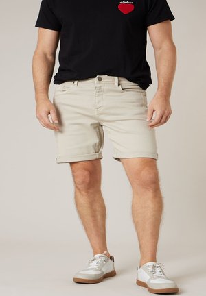 Homme portant un short beige retroussé, des baskets blanches et un t-shirt noir avec un cœur rouge et du texte sur la poitrine.
