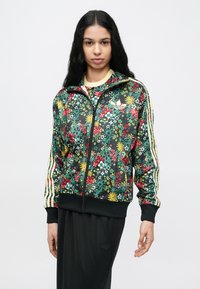 Frau trägt eine schwarze, mit Blumenmuster versehene Jacke mit Reißverschluss, gelben Streifen an den Ärmeln und schwarze Hose, steht vor einem schlichten weißen Hintergrund.