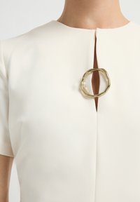 Close-up van een witte jurk met korte mouwen, voorzien van een keyhole-halslijn die is vastgemaakt met een onregelmatige gouden ronde sluiting op de borst.
