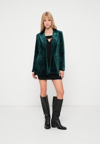 Σμαραγδένιο πράσινο σακάκι από βελούδο με λοξούς γιακάδες και δύο μπροστινές τσέπες, σε συνδυασμό με μαύρο φόρεμα και μαύρες μπότες έως το γόνατο.