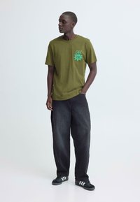 Olijfgroene T-shirt met korte mouwen en een graphic print op de borst, gecombineerd met zwarte wijde broekspijpen en zwarte sneakers met strepen.