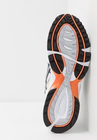 Sportssko med en vit mesh-överdela och svarta accenter, en texturerad grå mellanplatta, och en hållbar svart sula med orange detaljer.