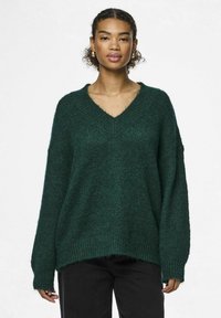 Zaļš adīts džemperis ar dziļu V veida dekolte, brīva piegriezuma un garām piedurknēm. Tekstūrēta auduma ar ribotām aprocēm un apakšmali. Kombinēts ar melniem biksēm.