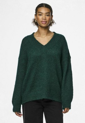 Grøn strikket sweater med dyb V-hals, løs pasform og lange ærmer. Tekstureret stof med ribstrikkede manchetter og kant. Sammen med sorte bukser.