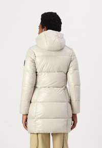 Lichtbeige pufferjas met een capuchon, voorzien van een glanzende afwerking, opgeblazen secties en een getailleerde taille, die tot halverwege de dijen reikt.