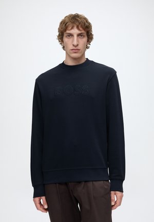 Jeune homme aux cheveux bouclés portant un sweat-shirt noir avec « BOSS » en relief sur la poitrine et un pantalon marron foncé sur un fond uni.