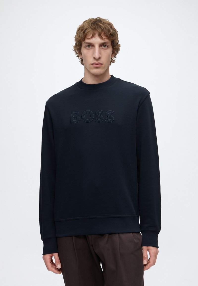 Jeune homme aux cheveux bouclés portant un sweat-shirt noir avec « BOSS » en relief sur la poitrine et un pantalon marron foncé sur un fond uni.