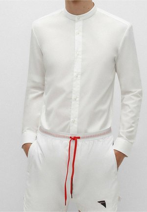 Mann trägt ein weißes Hemd mit Stehkragen und weiße Shorts mit rotem Kordelzug und Hugo-Logo-Patch, Hände in den Taschen.