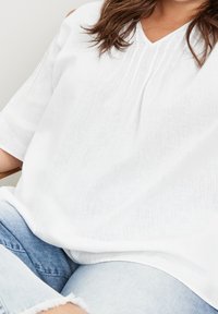 Femme portant un chemisier blanc ample à manches trois quarts et un jean bleu clair avec des ourlets effilochés, assise les jambes croisées.