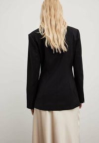 Blazer negro ajustado con un diseño estructurado, que presenta mangas largas, una silueta ceñida y puños abotonados, combinado con una falda color crema.