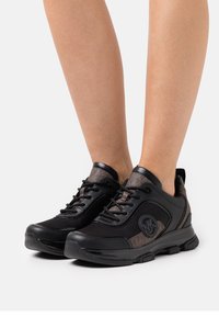 Chaussures de sport noires et brunes avec des panneaux en maille, des lacets ronds et un dos texturé. Présence d'un logo circulaire sur le côté.