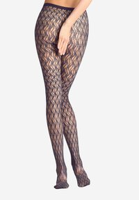 Femme portant des bas en dentelle noire transparente, se tenant les jambes croisées et une main posée sur la cuisse, sur fond blanc.