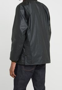 Černá lehká bunda s hladkou texturou, límcem a bočními kapsami. Obsahuje detaily se stiskacími knoflíky a je dokončena pleteným manžetem.