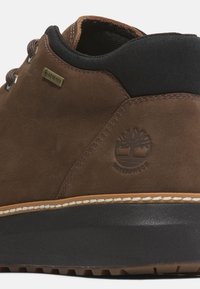 Zapato impermeable marrón con empeine de cuero liso, cuello negro, costuras blancas en contraste y suela de goma texturizada. Presenta el logo de Gore-Tex.