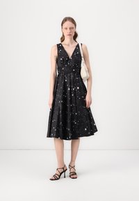Black dress with a V-neck, starry pattern, cinched waist, and flared skirt. Worn with strappy black heels and a small white bag.