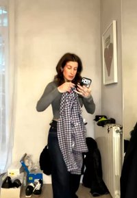 Femme aux cheveux foncés tenant un téléphone et une chemise à carreaux dans une pièce avec des chaussures, des vêtements et une œuvre d'art encadrée en forme de cœur sur le mur.
