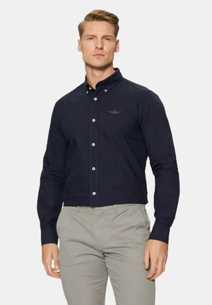 Camicia blu navy con maniche lunghe, dotata di colletto, bottoni bianchi e un discreto dettaglio con logo. Abbinata a pantaloni grigi chiari.