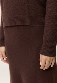 Hand vilar intill en mörkbrun stickad tröja och kjol med texturerade ärmar och ribbade fållar.