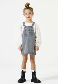 Denim overall jurk in lichtgrijs, met verstelbare bandjes, een voorkeer, gecombineerd met een wit blouse met lange mouwen en zwarte enkellaarzen.