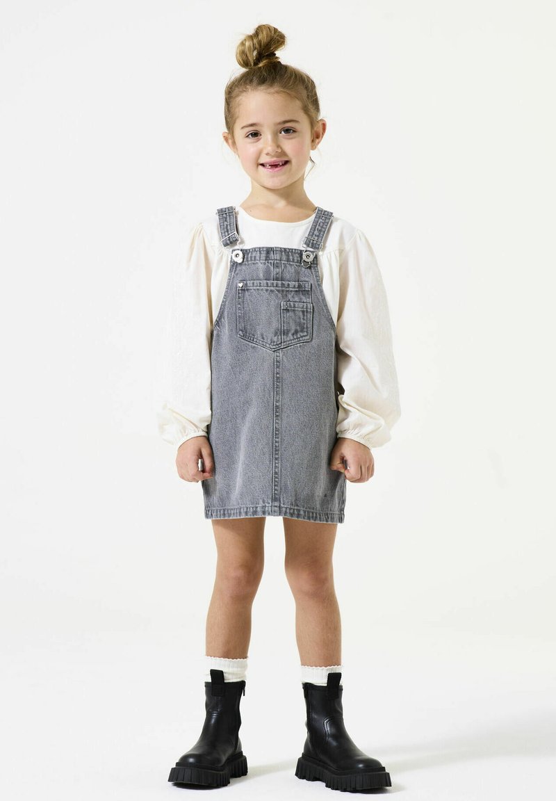 Denim overall jurk in lichtgrijs, met verstelbare bandjes, een voorkeer, gecombineerd met een wit blouse met lange mouwen en zwarte enkellaarzen.