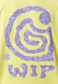 T-shirt jaune arborant un logo en spirale violet texturé et le texte « WIP » en bas. Le design présente un motif unique et irrégulier.