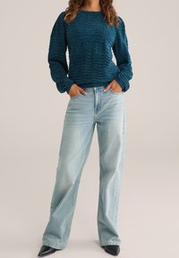 Personne portant un pull texturé bleu sarcelle foncé, un jean large bleu clair, et des chaussures noires à bout pointu, les mains dans les poches avant.