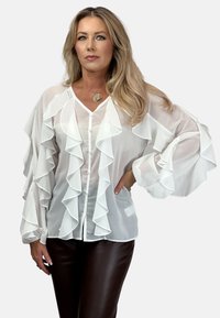 Biała, zwiewna bluzka z falbanami z przodu i przy rękawach, z dekoltem w kształcie litery V i zapięciem na guziki. Gładka tekstura, lekki materiał.
