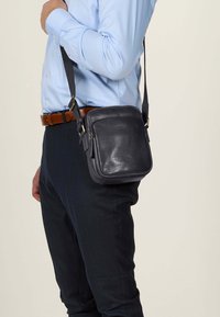 Borsa a tracolla in pelle nera con forma squadrata, accenti in oro e chiusura con zip, indossata sopra una camicia azzurro chiaro e pantaloni blu navy.
