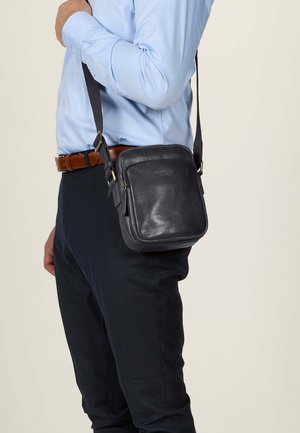 Borsa a tracolla in pelle nera con forma squadrata, accenti in oro e chiusura con zip, indossata sopra una camicia azzurro chiaro e pantaloni blu navy.